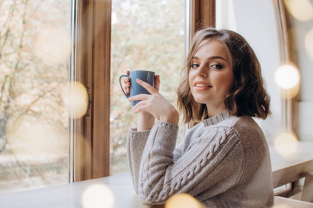 Piękna dziewczyna w swetrze pije herbatę lub kawę na tle okna i świateł