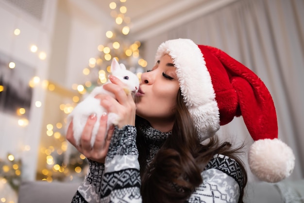 Piękna dziewczyna w świątecznym swetrze i czerwonym kapeluszu Świętego Mikołaja trzyma w dłoniach małego białego królika i całuje go Obchody świąt noworocznych