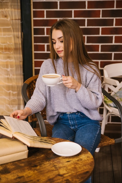 Piękna dziewczyna w szarym swetrze z dzianiny pije kawę i czyta książkę w przytulnej kawiarni, koncepcja przyjemnego wypoczynku i komunikacji. Student lub bizneswoman.