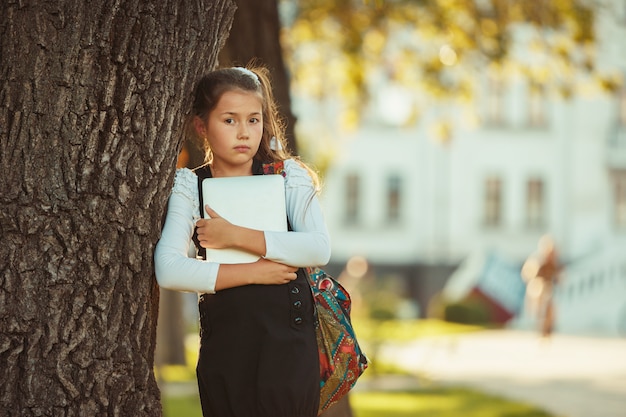 Piękna dziewczyna w wieku szkolnym stoi przy drzewie i trzyma tabletkę. Uczennica ubrana w sukienkę