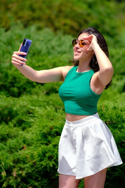 Piękna dziewczyna w zielonym topie i białych szortach spaceruje po parku miejskim w słoneczny letni dzień Robiąc selfie śmiejąc się trzymając telefon komórkowy