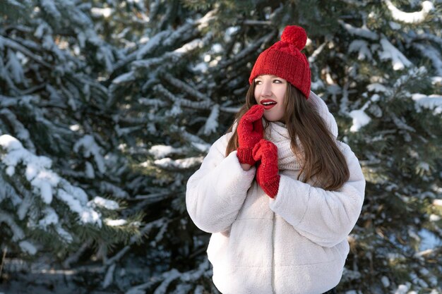 Piękna dziewczyna z czerwonymi ustami w czerwonym kapeluszu i mitynkach w zimowym lesie Uśmiechnięta młoda kobieta na spacerze