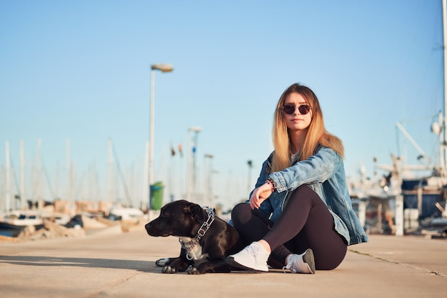 Piękna dziewczyna z jej ciemnym psem siedzą w portowej dżinsowej kurtce, letni dzień na zewnątrz w porcie