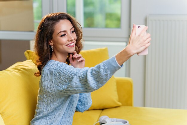 Piękna Dziewczyna Z ślicznym Uśmiechem Robi Selfie Na Tle Kuchni. Wesoła Dziewczyna Rozmawia Online Ze Swoim Chłopakiem Za Pomocą Smartfona.