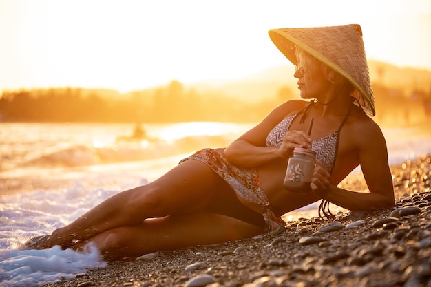 Zdjęcie piękna dziewczyna z smoothie nad morzem w słomkowym azjatyckim trójkątnym kapeluszu