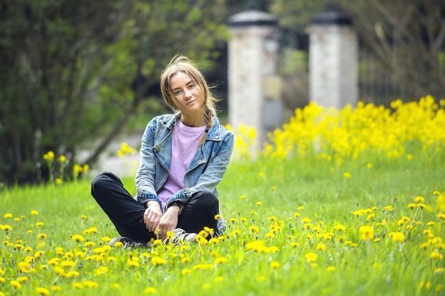 Piękna dziewczyna, z włosami zaplecionymi w warkocz, siedzi na jasnozielonym trawniku z żółtymi kwiatami