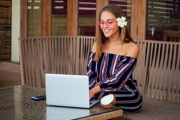 Piękna i młoda biznesowa kobieta w stylowej bluzce w paski z odkrytymi ramionami, siedząca przy stole z laptopem na tarasie letniej restauracji nad oceanemfreelancer dziewczyna pracująca online