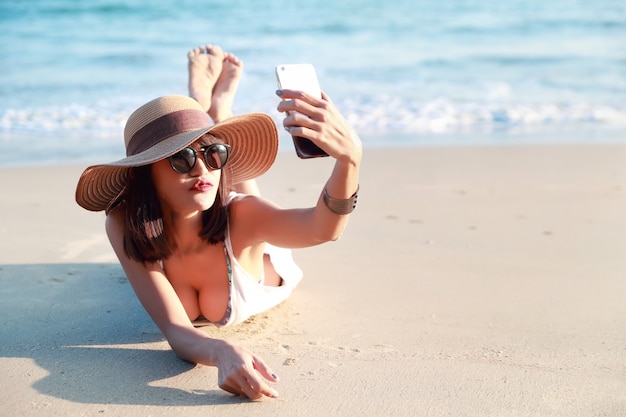 Piękna i seksowna kobieta korzysta z telefonu komórkowego podczas wakacji robi autoportret i całuje się z aparatem na plaży