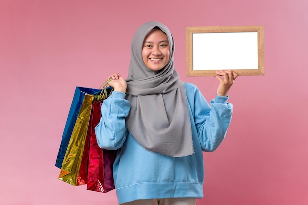 Zdjęcie piękna indonezyjska kobieta trzymająca papierowe torby na zakupy i pokazująca małą pustą tablicę na znaki reklamowe lub miejsce na kopię