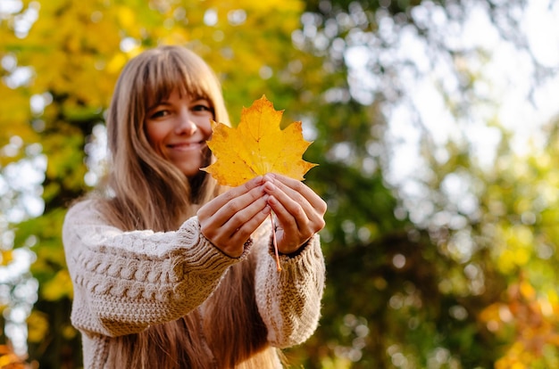 Piękna jesienna kobieta stojąca w pobliżu kolorowych jesiennych liści Całkiem szczęśliwy wesoły model patrząc na kamerę Młoda kobieta z jesiennym liściem w dłoni jesienny nastrój relaks chodzenie w przyrodzie