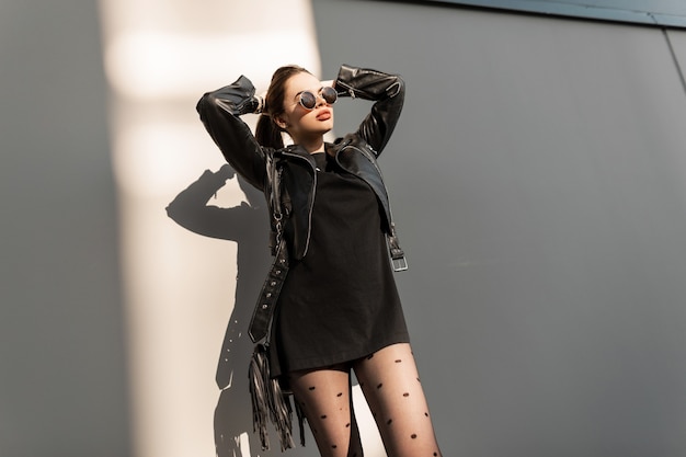 Piękna kobieta hipster z stylowe okrągłe okulary w czarną skórzaną kurtkę moda i sukienka z sexy rajstopy stawia przy ścianie w słońcu na ulicy. Miejski kobiecy styl i moda