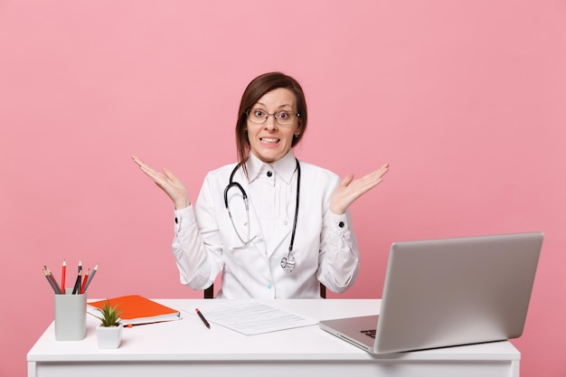 Piękna kobieta lekarz siedzi przy biurku pracuje na komputerze z dokumentem medycznym w szpitalu na białym tle na pastelowym różowym tle ściany. Kobieta w medycznym stetoskopie toga okularów. Koncepcja medycyny opieki zdrowotnej