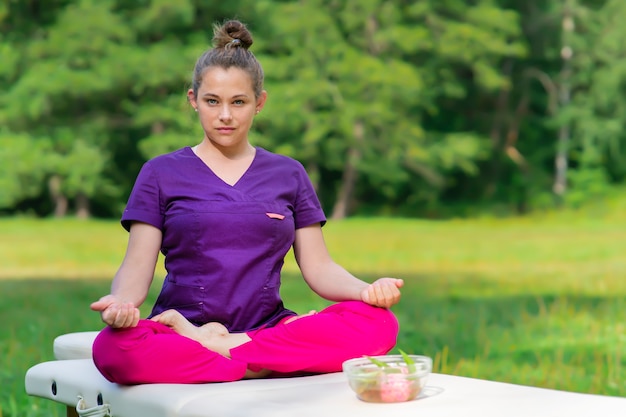 Piękna kobieta masażysta profesjonalny terapeuta w mundurze, masażysta siedzi na stole do masażu w pozycji lotosu na zewnątrz w słoneczny letni park. Opieka zdrowotna, koncepcja masażu