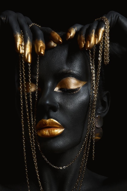 Piękna kobieta pomalowana na czarny kolor skóry body art, złoty łańcuszek w dłoniach i wokół szyi. Złoty makijaż usta powieki, opuszki palców paznokcie w złotym kolorze farbą. Profesjonalny makijaż