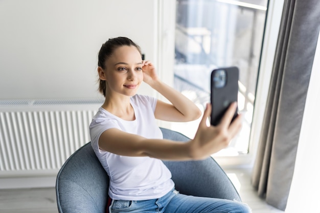 Piękna kobieta robi selfie swoim smartfonem i uśmiecha się siedząc w dużym krześle w domu