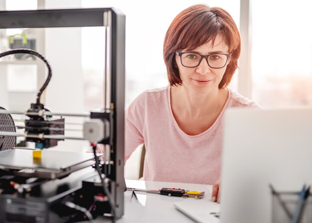 Piękna kobieta siedzi i patrzy na laptopa i pracuje nad projektem z nowoczesną drukarką 3D