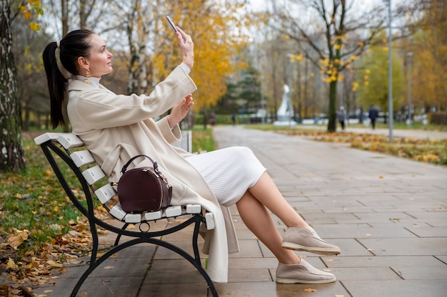 piękna kobieta siedzi na ławce w parku... kobieta w średnim wieku robi selfie przez telefon w parku