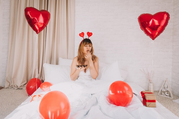 Piękna kobieta siedzi w łóżku z białą pościelą i je czekoladki. Walentynki.