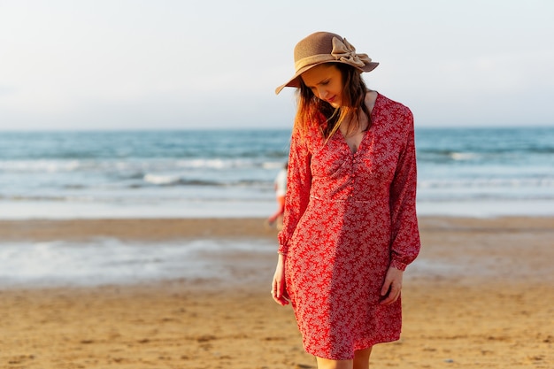 Piękna Kobieta Spaceru Na Plaży Wiosną Kapelusz I Czerwona Sukienka. Koncepcja Wakacje Wielkanocne. Hiszpania