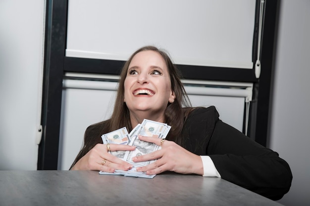 Piękna kobieta trzymająca pieniądze uśmiecha się i przytula dużo pieniędzy Uśmiechnięta kaukaska bizneswoman w czarnym garniturze uśmiecha się mocno zębami do kamery w biurze Szczęśliwa Żydówka przytula dolary