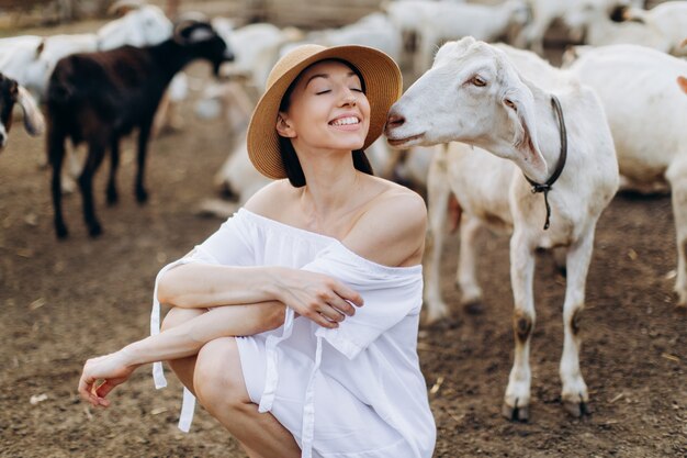 Piękna kobieta w białej sukni i beżowym kapeluszu wśród kóz na ekologicznej farmie