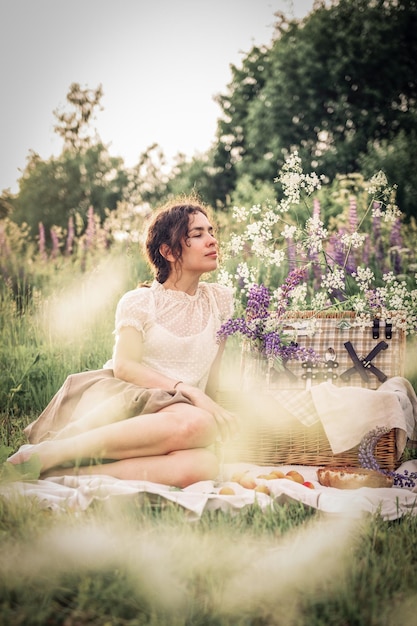 Piękna kobieta w białej sukni siedzi na trawie z koszem śniadaniowym