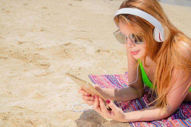 Piękna kobieta w bikini i kapeluszu śpi na plaży, słucha piosenki z tabletem