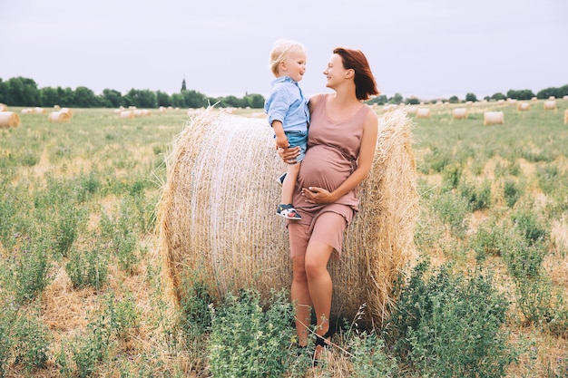 Piękna kobieta w ciąży i dziecko bawią się na polu pszenicy ze stogami siana w letni dzień na łonie natury