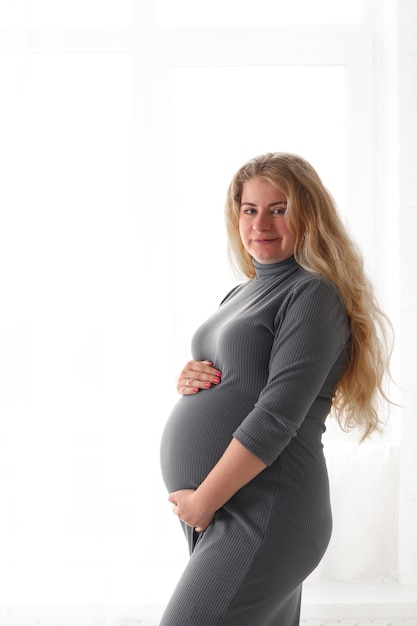 Zdjęcie piękna kobieta w ciąży stoi w pobliżu okna trzymając brzuch i patrząc na kamerę selective focus copy space koncepcja ciąży i macierzyństwa