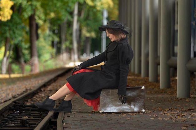 Piękna kobieta w czarnym płaszczu i kapeluszu siedzi na skórzanej walizce, czekając na pociąg na stacji Zdjęcie w stylu lat 3040