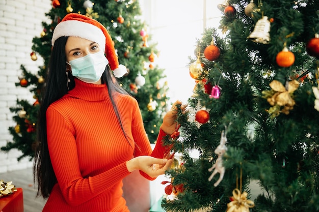 Piękna kobieta w czerwonej sukience i czapce Mikołaja z maską chirurgiczną siedzi otoczona kolorowymi pudełkami z prezentami i cieszy się samotnie Świętami Bożego Narodzenia w domu.