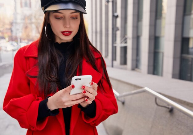 Piękna kobieta w czerwonym płaszczu i czarnym kapeluszu wysyła SMS-y na swoim telefonie na ulicy