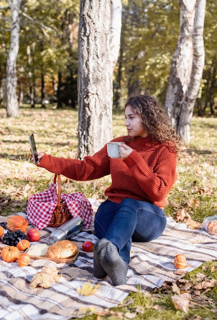 Piękna kobieta w czerwonym swetrze na pikniku w jesiennym lesie za pomocą telefonu komórkowego