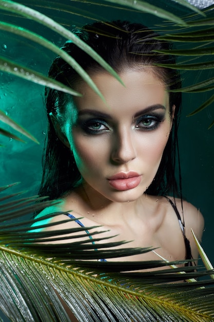 Piękna kobieta w dłoni pozostawia mokry makijaż, tropikalny portret dziewczyny w zielony strój kąpielowy w gałęzi palmy w studio, dym i krople deszczu na szkle. Seksowna kobieta z jasnozielonym makijażem