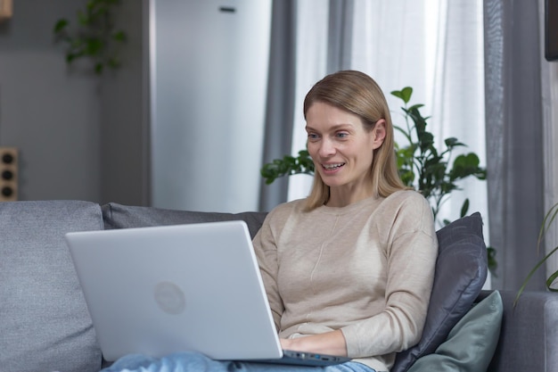 Piękna kobieta w domu siedzi na kanapie, uśmiechając się i pracując na laptopie