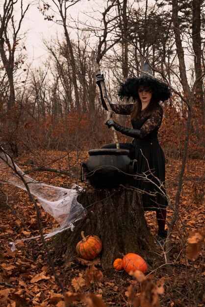 Piękna kobieta w kapeluszu czarownic i kostium przygotowuje miksturę w kotle w jesiennym lesie Koncepcja Halloween Selektywne skupienie