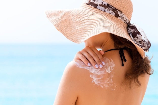 Piękna kobieta w kapeluszu nakłada krem przeciwsłoneczny na ramię na plaży