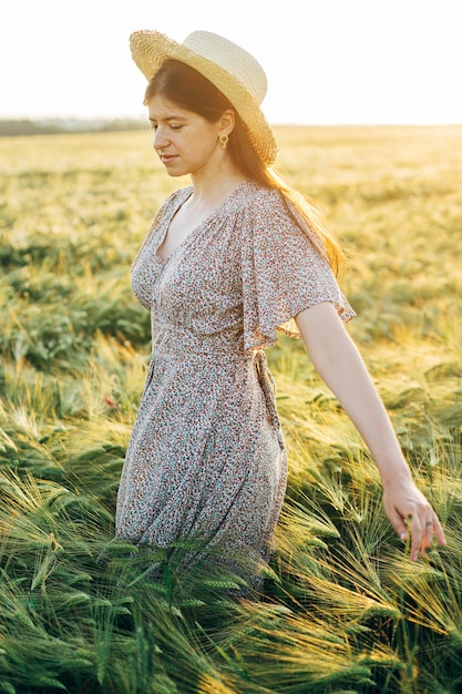 Piękna kobieta w kwiecistej sukience spacerująca po polu jęczmienia w świetle zachodzącego słońca Stylowa kobieta w słomkowym kapeluszu i relaksująca się wieczorem na letniej wsi Atmosferyczny moment rustykalne powolne życie