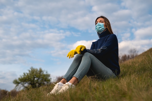 Piękna kobieta w masce medycznej i rękawiczkach siedząca na zielonym wzgórzu