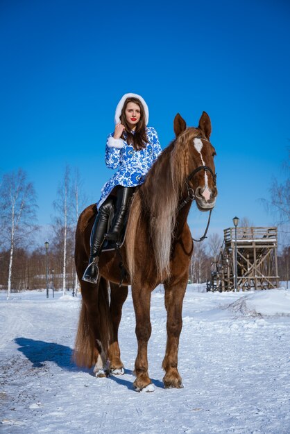 Piękna kobieta w niebieskim płaszczu na koniu zimą w słoneczny dzień