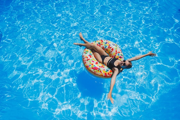 Piękna kobieta w okularach przeciwsłonecznych w basenie unosi się na dmuchanym kółku do pływania w czarnym stroju kąpielowym letnie zdjęcie fotografia pływania letnie zdjęcia kobiety