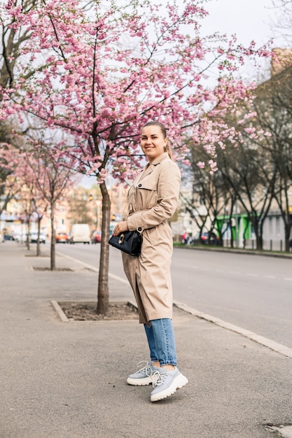 Piękna Kobieta W Pobliżu Drzew Sakura. Różowe Kwiaty Kwitną Na Drzewach Na Ulicy Miasta. Wokół Kwitną Drzewa. Koncepcja Wiosennego Czasu