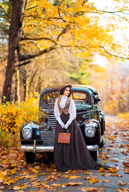 Piękna kobieta w sukni vintage, koronkowej bluzce i kapeluszu z welonem stojąca w pobliżu retro brązowego samochodu