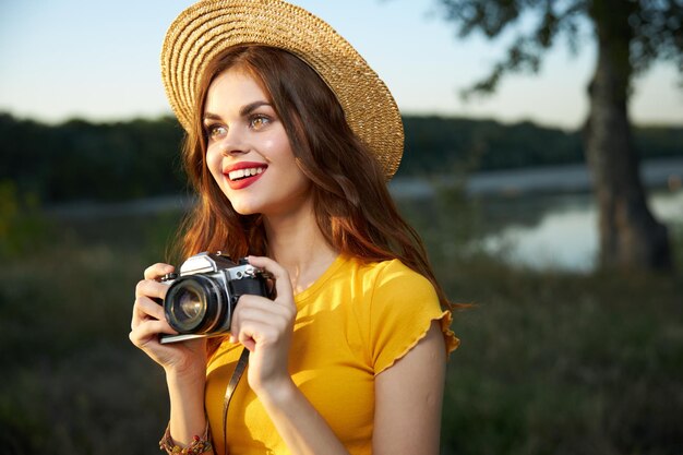 Piękna kobieta z aparatem fotograficznym w rękach na przyrodzie nosząca kapelusz czerwone usta uśmiech patrzy na zdjęcie wysokiej jakości