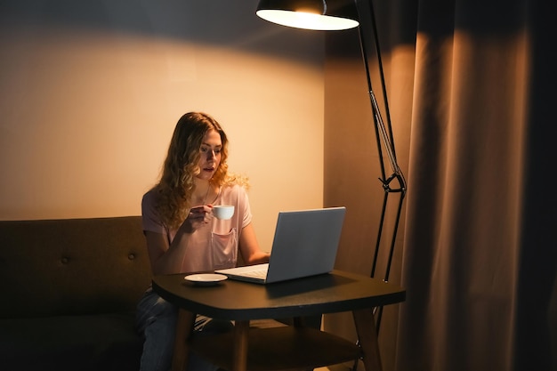 Zdjęcie piękna kobieta z filiżanką kawy pracuje wieczorem na laptopie dziewczyna z notebookiem w ciemnym pokoju di