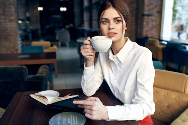 Zdjęcie piękna kobieta z filiżanką w ręku siedzi przy stole w kawiarni i książkę w ręku model restauracji wysokiej jakości zdjęcie