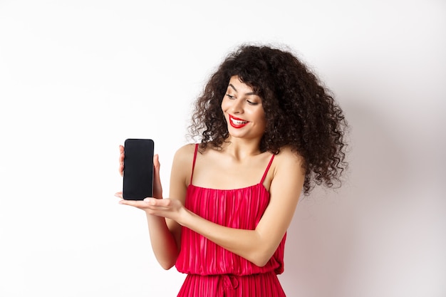 Piękna kobieta z makijażem i kręconymi włosami, pokazując pusty ekran smartfona, demonstrując aplikację, stojąc na białym tle.