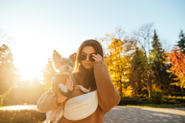 Piękna kobieta z psem na rękach w parku na tle zachodu słońca pozuje do kamery