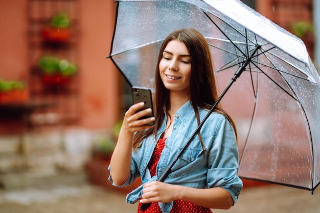 Piękna kobieta za pomocą smartfona na ulicy w deszczową pogodę uśmiecha się trzymając parasol Letni deszcz