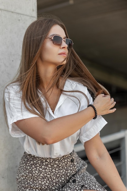 Piękna kobieta ze stylowymi, zabytkowymi okularami przeciwsłonecznymi w modnych ubraniach z białą koszulą w mieście